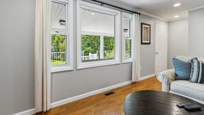 Une pièce avec un mur gris, une porte blanche, un canapé gris agrémenté de coussins bleus et blancs, une table basse noire avec des télécommandes, et une fenêtre avec des rideaux blancs.