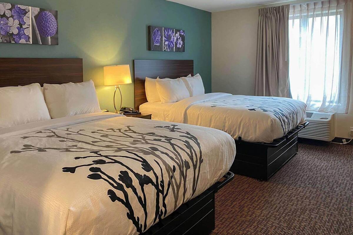 A hotel room with two beds, a nightstand with a lamp, a wall-mounted artwork, and a window with curtains.