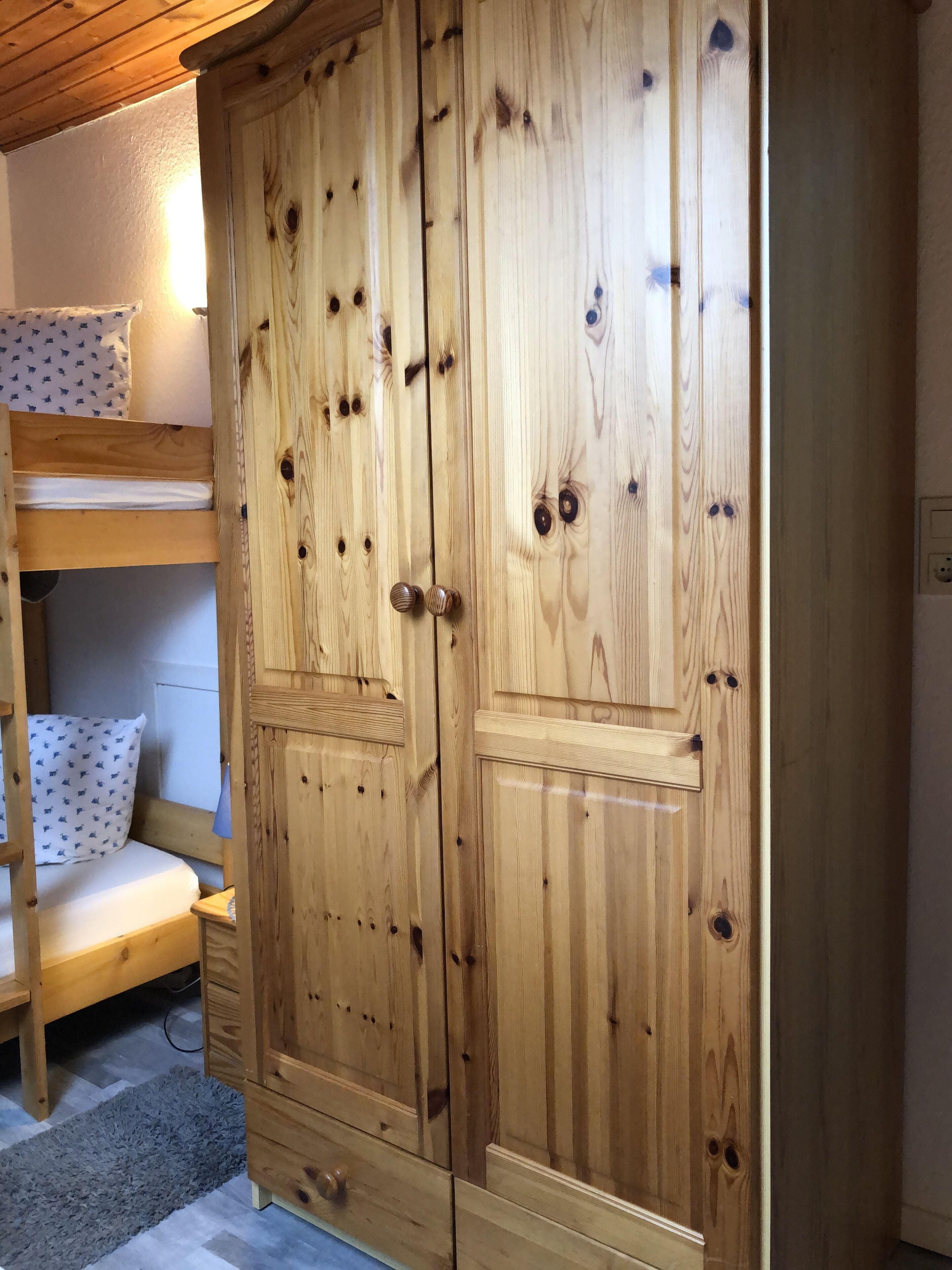 A wooden wardrobe with two doors, a bunk bed with a patterned pillow, and a small bedside table.