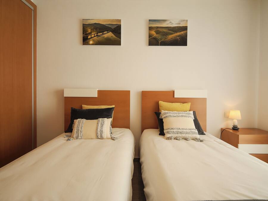 A hotel room with two beds, a wooden wardrobe, a nightstand with a lamp, and two landscape paintings on the wall.