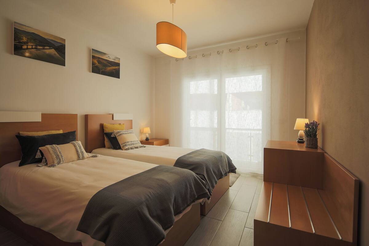 A hotel room with two beds, a bedside table with a lamp, a small bench, and artwork on the wall.