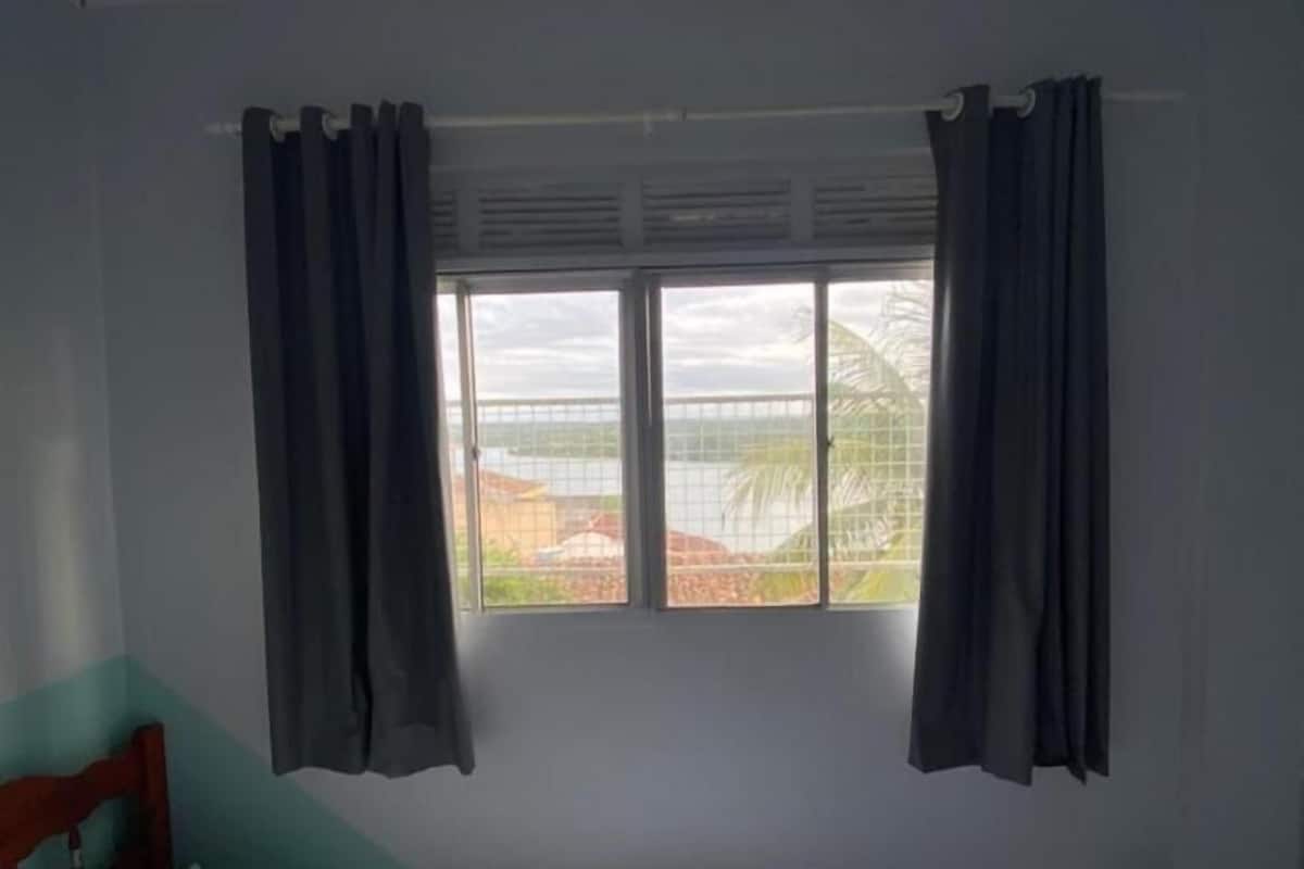 A single bed with a white sheet, two grey curtains, and a window with a view of greenery and a fence.