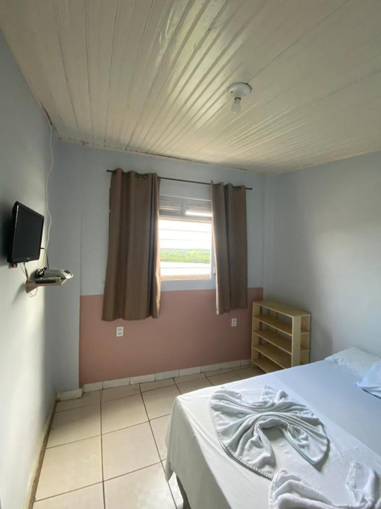 Ein Schlafzimmer mit einem Bett, einem an der Wand montierten Fernseher, einem Fenster mit Vorhängen, einer Holzablage und einem Deckenventilator.