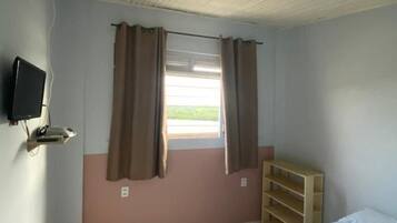 Ein Schlafzimmer mit einem Bett, einem an der Wand montierten Fernseher, einem Fenster mit Vorhängen, einer Holzablage und einem Deckenventilator.