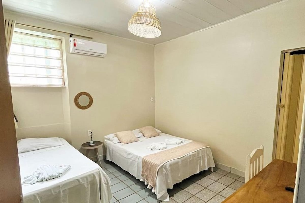 A room with two beds, a wooden table, a ceiling fan, and a window with blinds.