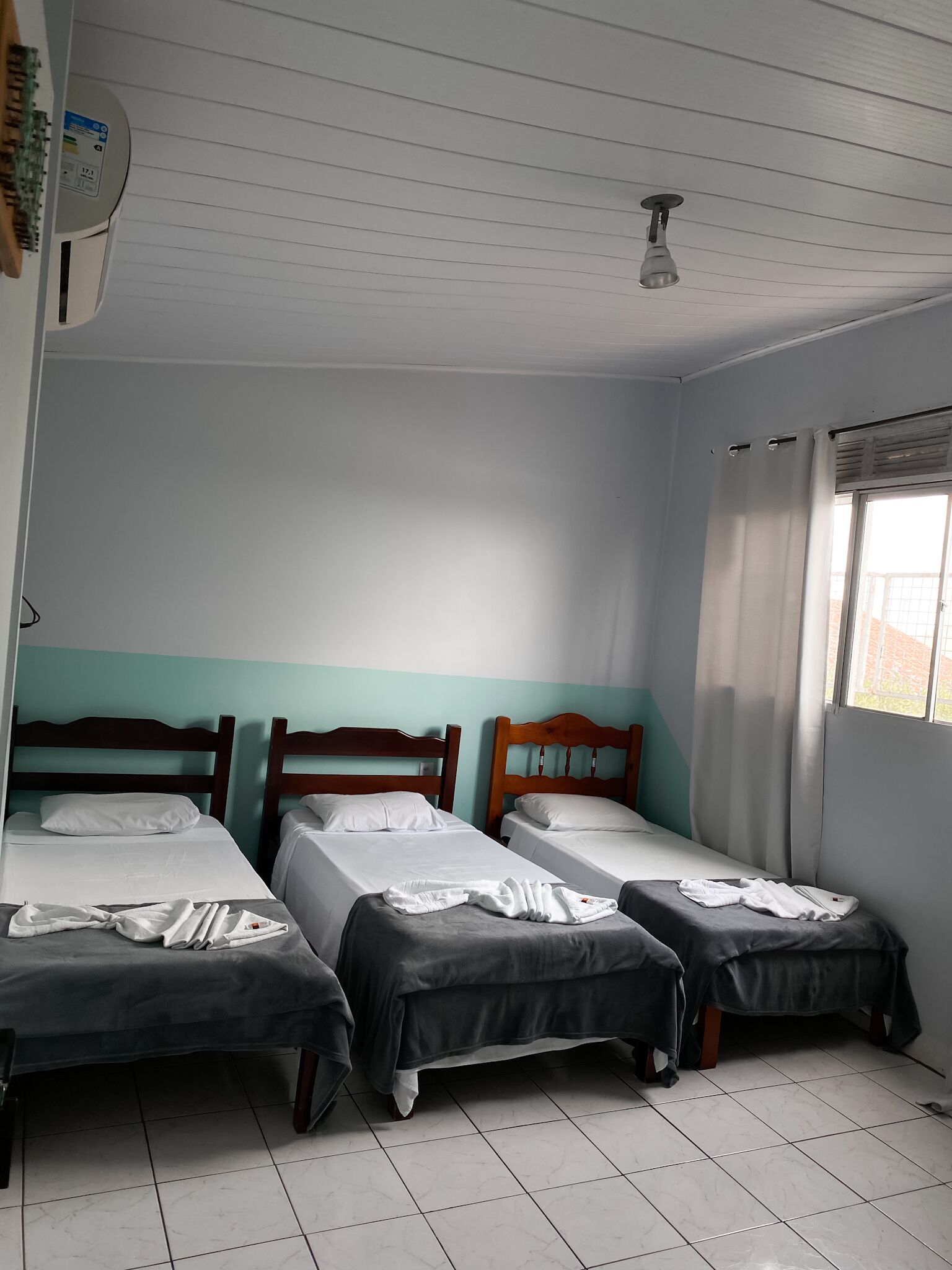 A room with three single beds, a ceiling fan, a window with curtains, and a wall-mounted air conditioner.