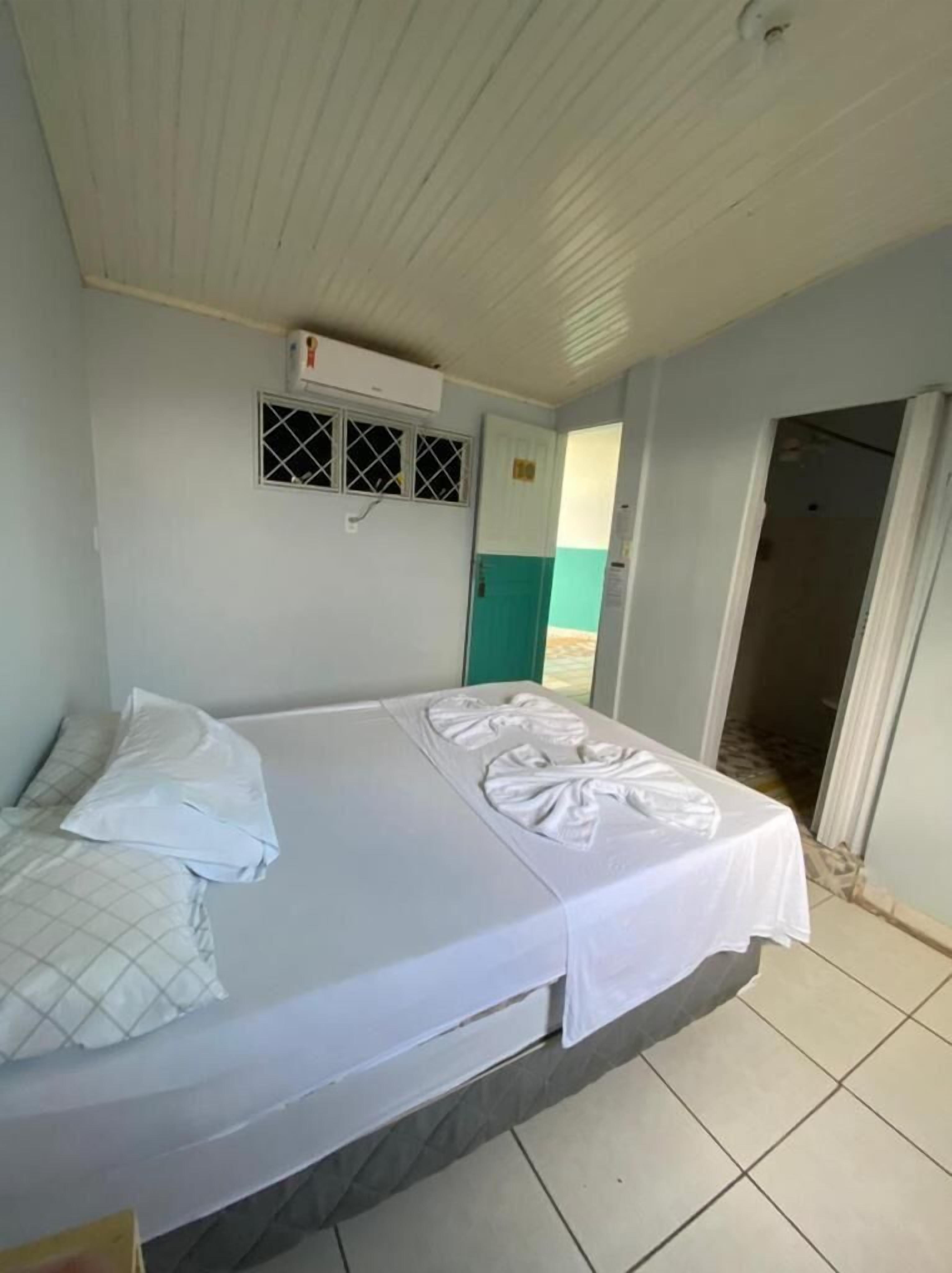 A single bed with a white bedspread and pillows, a window with a metal grille, and a door leading to another room.