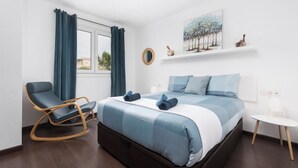 Un dormitorio moderno con cama, una mecedora, una mesita y una ventana con cortinas.
