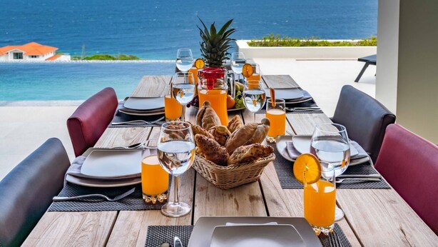 A table set for a meal with a view of the ocean, including plates, glasses, and a basket of bread. - Stunning Luxury Villa/ Private Infinity Pool with Panoramic Sea Views (Sint Willibrordus)