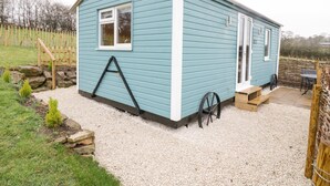 A small, light blue mobile home with a white door, a small porch, and a wheelbarrow. - Robins Nest (Southam)