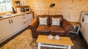 A cozy cabin-style room with a brown leather sofa, a wooden table with two cups, a kitchen area with a microwave, and a window with a view of the outdoors. - Robins Nest (Southam)
