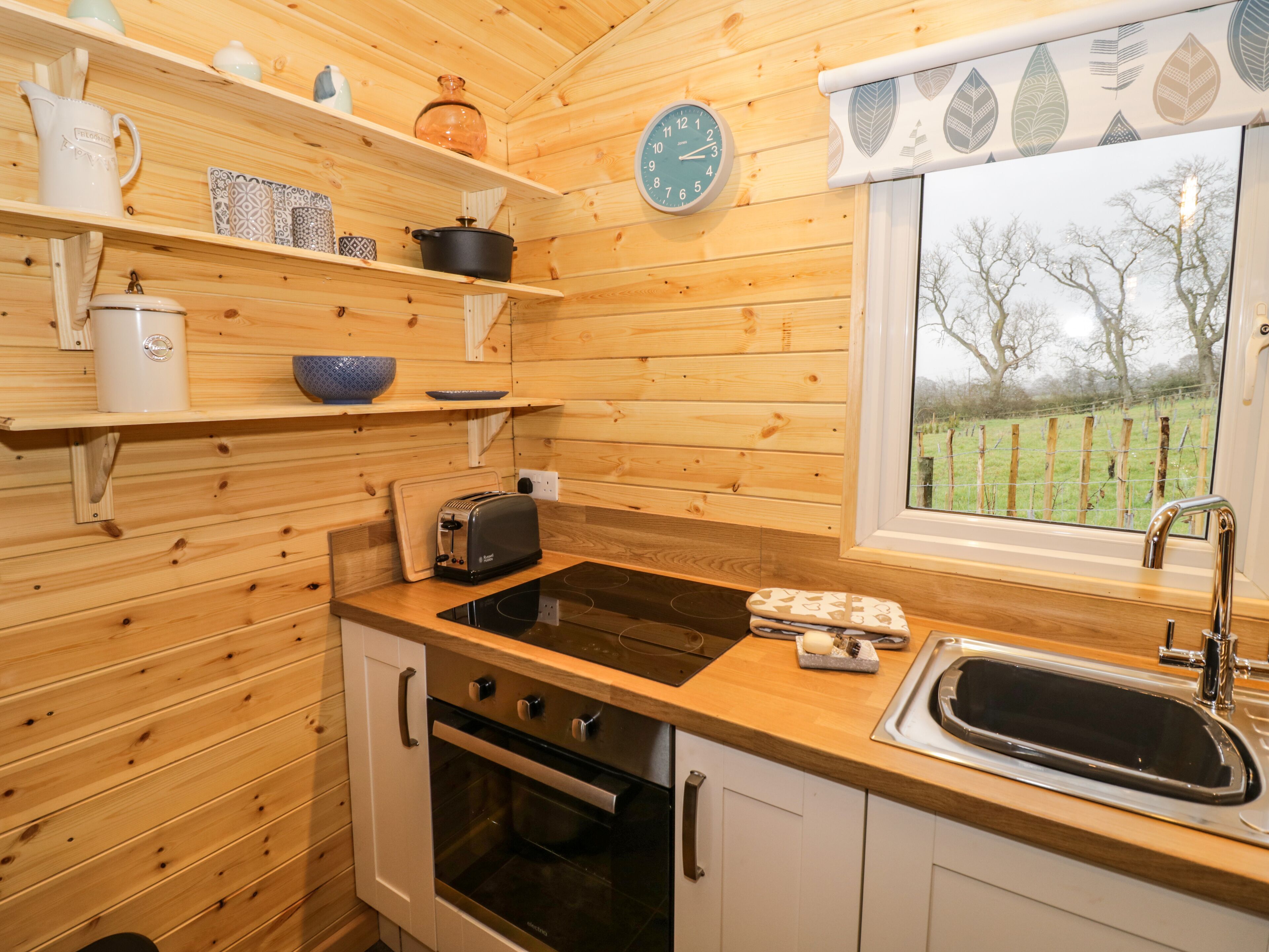 A compact kitchen with wooden walls, a built-in oven, a sink, and a window with a view of a grassy field.