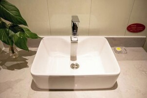 A white rectangular bathroom sink with a chrome faucet, a small yellow soap dish, and a potted plant to the left.