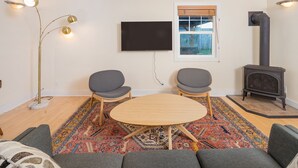 Una sala de estar con una mesa redonda de madera, dos sillas grises, un sofá, una lámpara de pie, un televisor de pantalla plana, una ventana con persianas y un estufa de leña.
