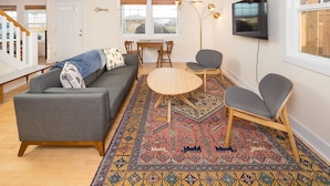 Una sala de estar moderna con un sofá gris, una mesa de centro redonda de madera y dos sillones grises. Una alfombra colorida cubre el suelo, y un televisor está montado en la pared.