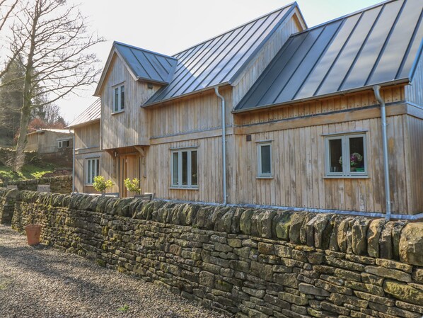A wooden house with a stone wall in front, metal roof, and multiple windows. - Oak Cottage (Rochdale)