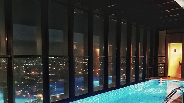 Una piscina interna moderna con vista sulla città, ampie finestre e pavimento in legno.
