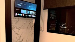 Una camera d'albergo con parete in marmo, TV a parete, finestra vista città e asciugamano piegato sul letto.
