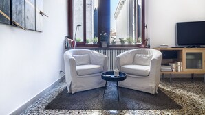 A compact living room with two white armchairs, a small coffee table, a TV on a wooden stand, and a window with a view of buildings. - ACQUARELLO, SPACIOUS APARTMENT IN THE HEART OF VENICE (Venise)