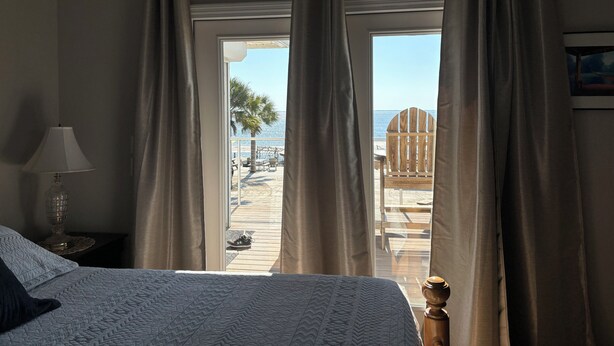A bedroom with a bed, a nightstand with a lamp, and a view of a beach and palm trees through the window. - Reel Em Inn Bay Front House With boat ramp (port saint joe/simmons bayou)