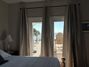 Une chambre à coucher avec un lit, une table de chevet avec une lampe, et une vue sur une plage et des palmiers par la fenêtre.