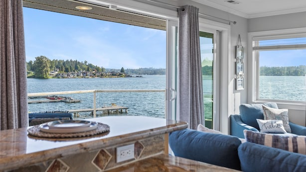 A room with a view of a lake and dock, a blue sofa, and a wooden table with a placemat. - Relaxing 4 bed, 4 bath, + private studio lakefront home on Lake Stevens! (Lake Stevens)