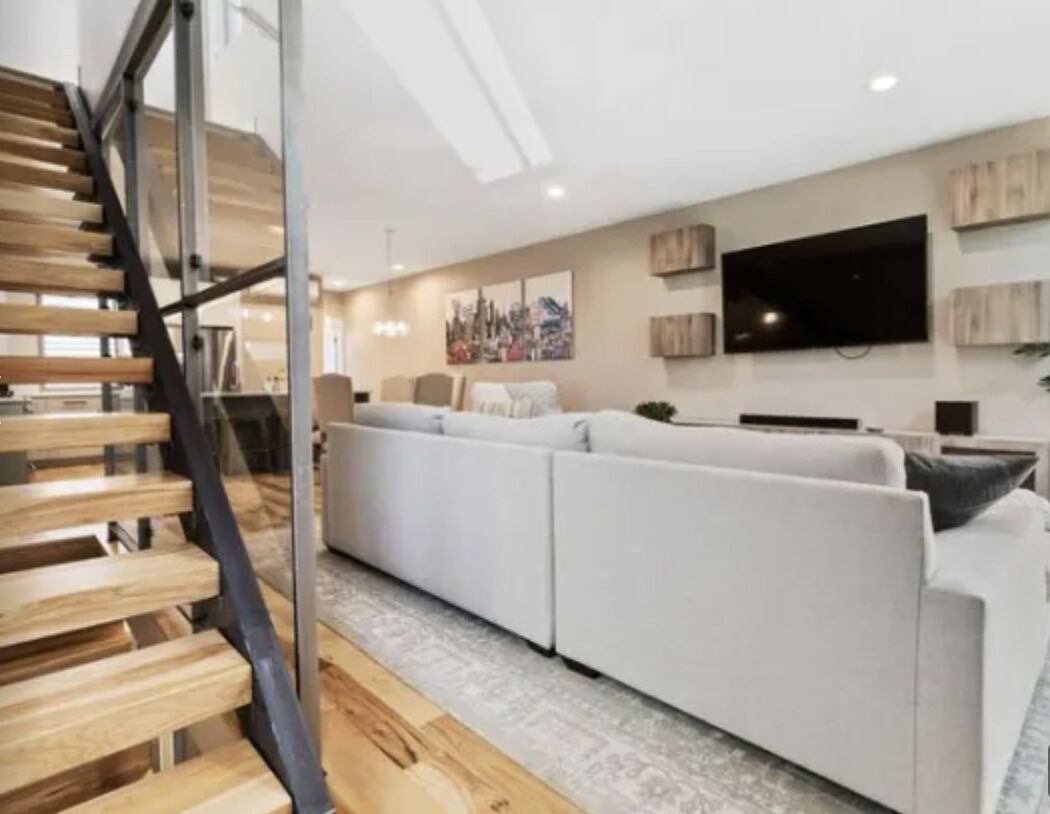A modern living room with a sofa set, a flat-screen TV mounted on the wall, and a wooden staircase.