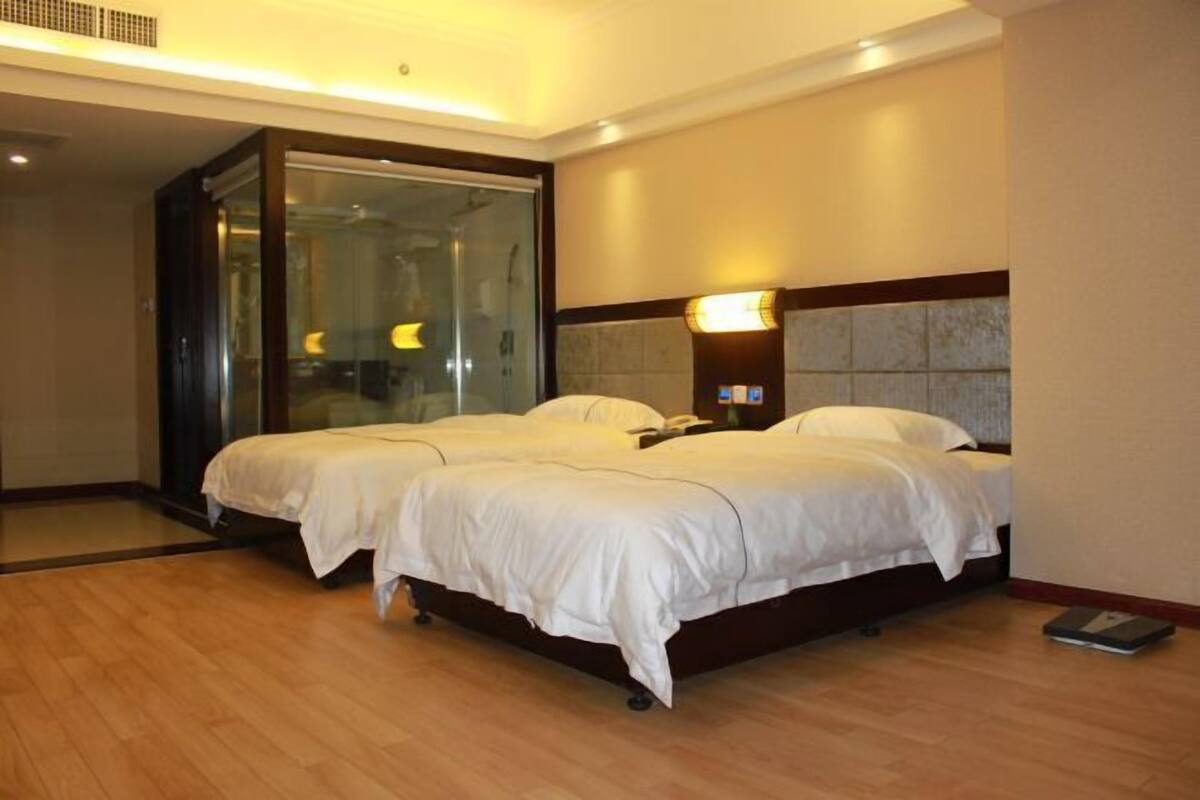 A hotel room with two beds, a glass shower enclosure, a nightstand with a lamp, and a wooden floor.