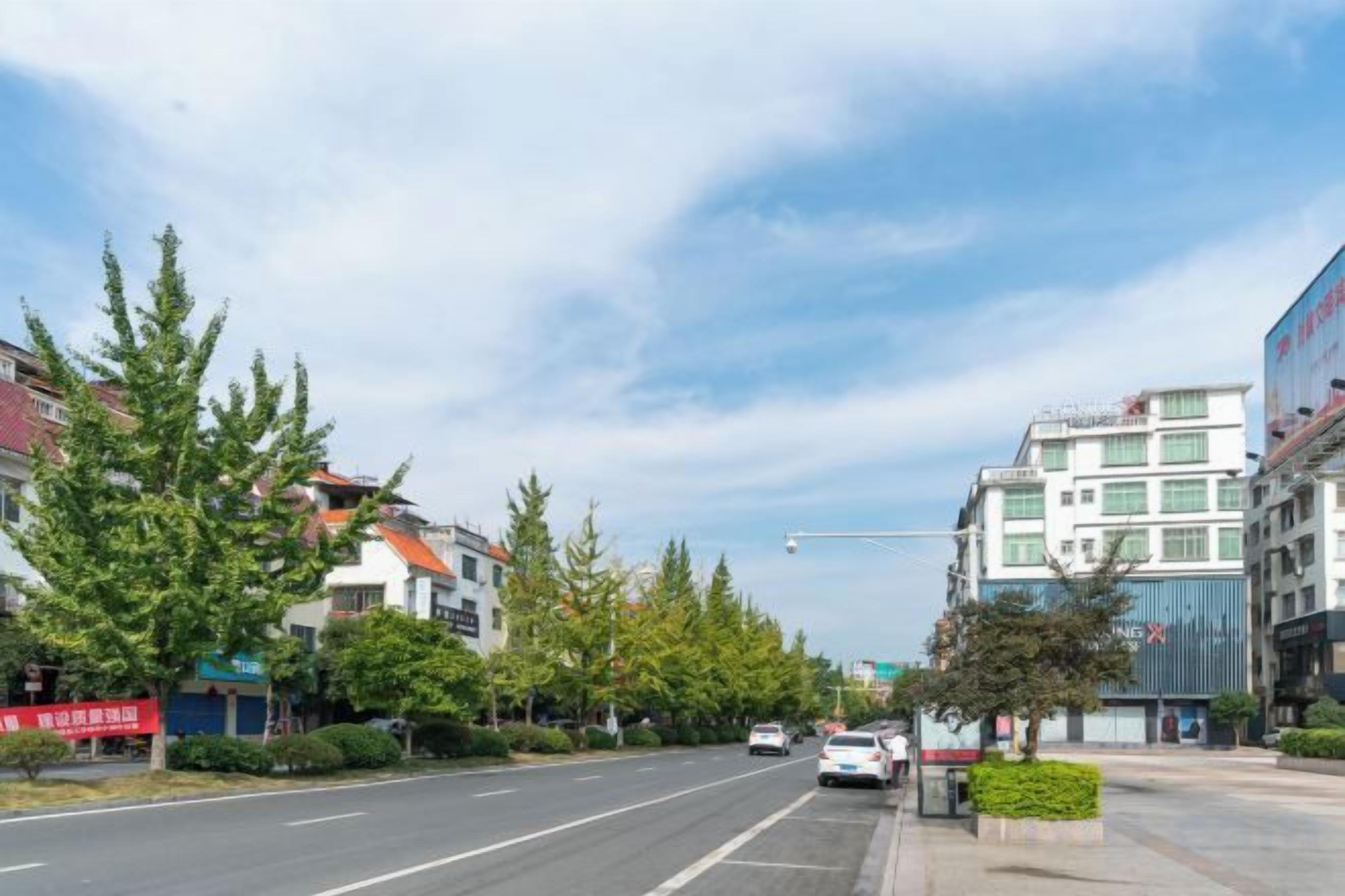 A wide urban street with modern buildings, a clear sky, and a few cars on the road.