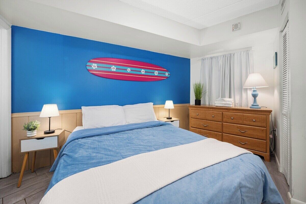 Une chambre d’hôtel comprenant un lit, deux tables de chevet, une commode et un mur décoré avec un motif de planche de surf.