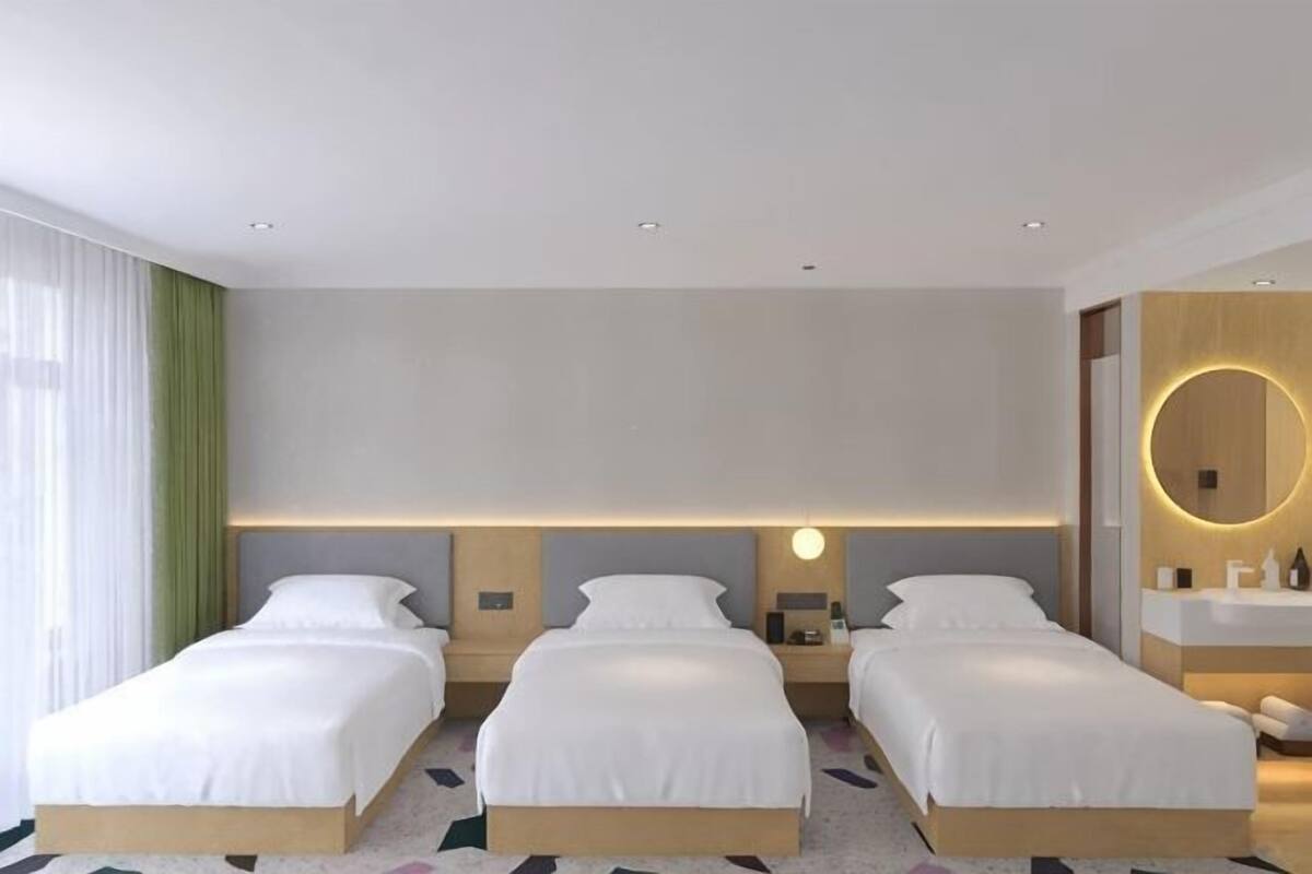 Moderni hotellihuone, jossa on kolme sänkyä, suuri peili ja neutraali värimaailma.
