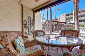 A round glass table with wicker chairs and cushions, a textured wall, and a view of palm trees and buildings.