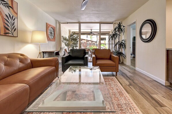 A living room with a brown leather sofa, a black armchair, a glass coffee table, and a patterned rug. There is a large window with a view of the outdoors, a ceiling fan, and a wall-mounted clock.