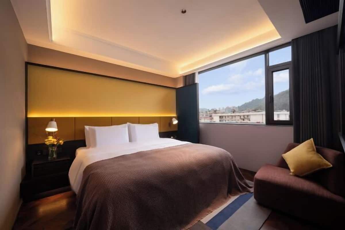 Een moderne hotelkamer met een groot bed, een nachtkastje met lamp, een bank met kussen en uitzicht op de stad door grote ramen.