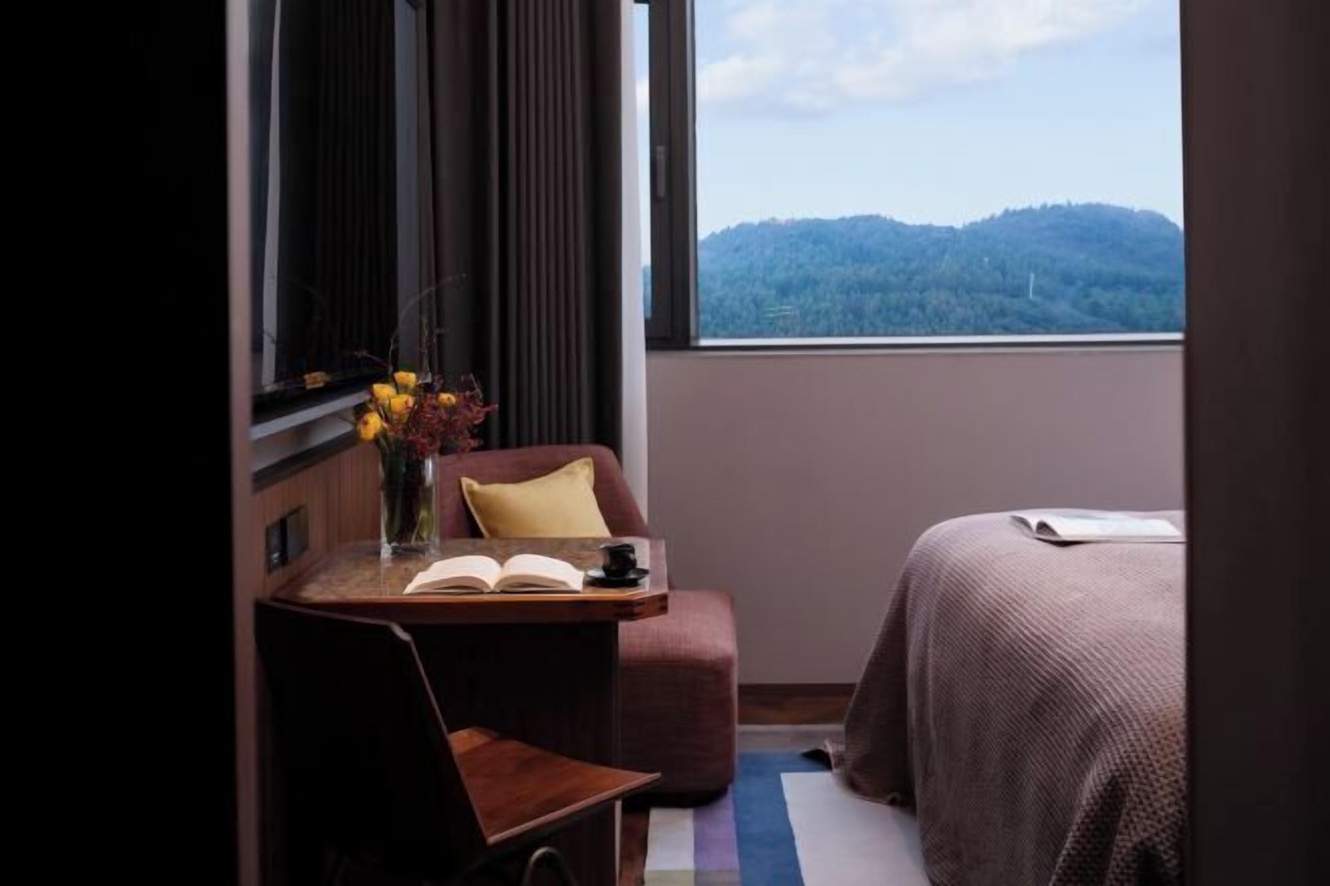 Un dormitorio con una cama, una mesita de noche con un libro y un florero, una ventana que da a una colina boscosa y una silla.