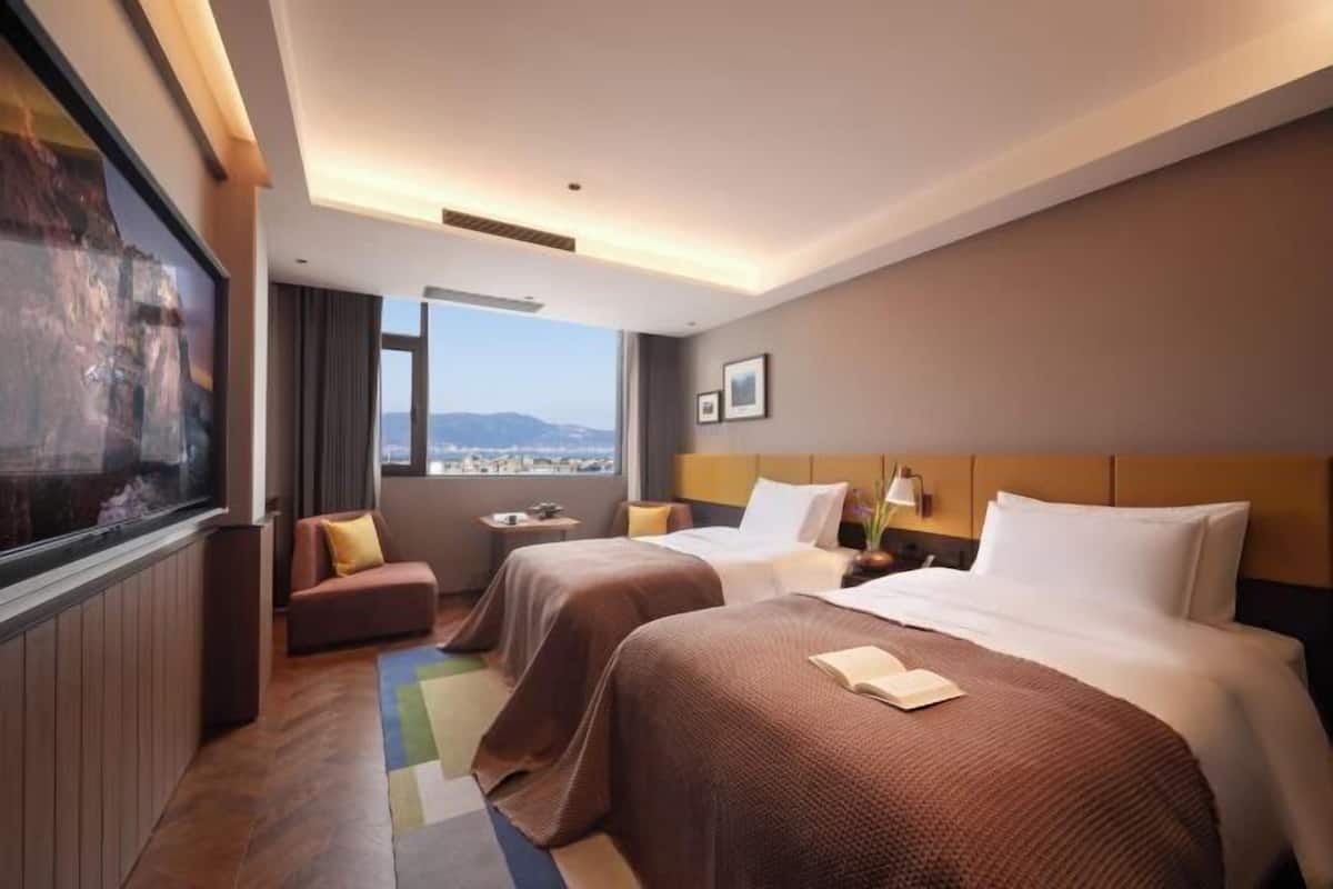 Een hotelkamer met twee bedden, een groot raam, uitzicht over de stad en een schilderij aan de muur.