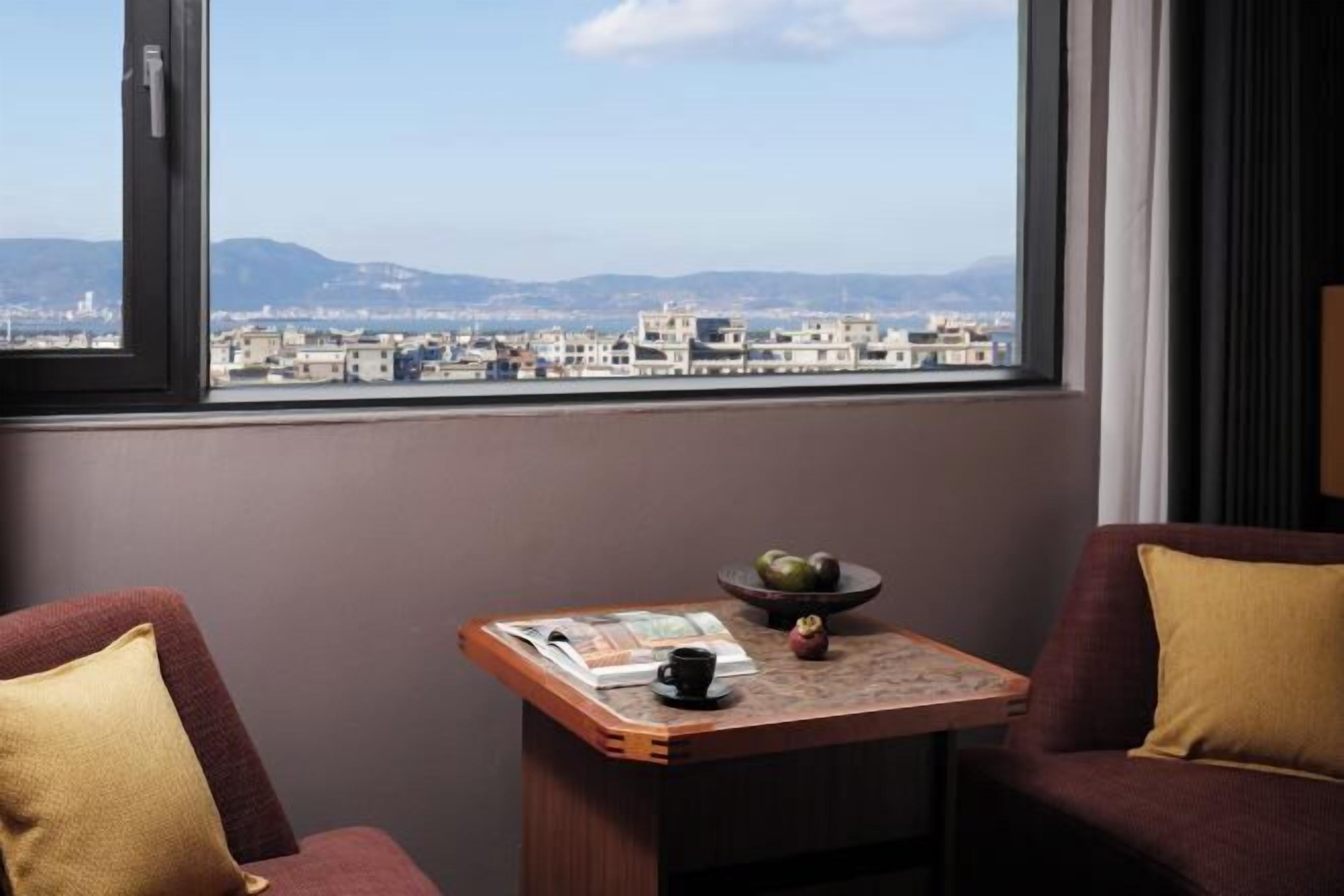 A room with a view of a cityscape through a window, a small table with a bowl of fruit, and two sofas with cushions.