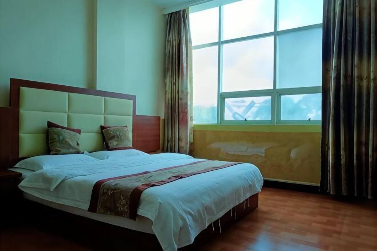 A neatly made bed with a headboard, a window with curtains, and a wooden floor.