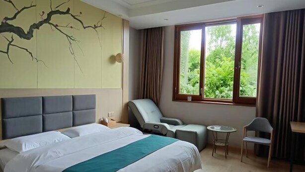 A hotel room with a large bed, a massage chair, a small table, and a view of greenery through the window. - Yintai Hotel (Menglian)