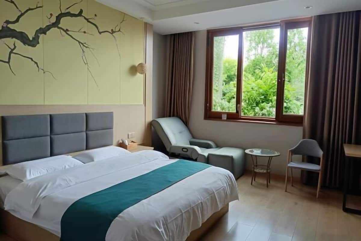 A hotel room with a large bed, a massage chair, a small table, and a view of greenery through the window.