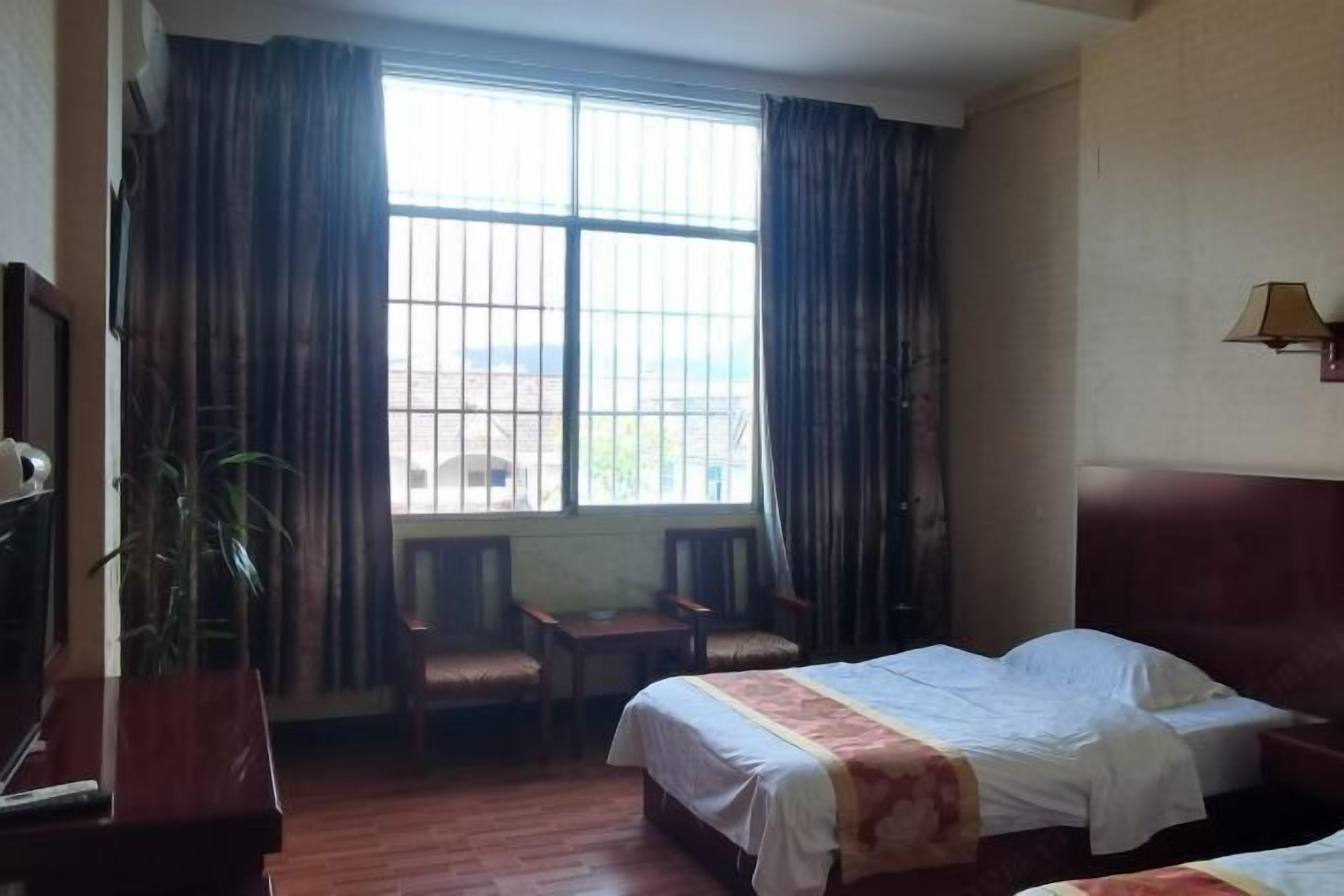 A hotel room with two beds, a desk, chairs, a potted plant, and a window with curtains.
