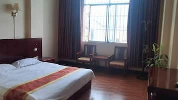 A hotel room with a bed, a desk, two chairs, a potted plant, and a window with curtains.