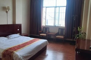 A hotel room with a bed, a desk, two chairs, a potted plant, and a window with curtains. - Yintai Hotel (Menglian)