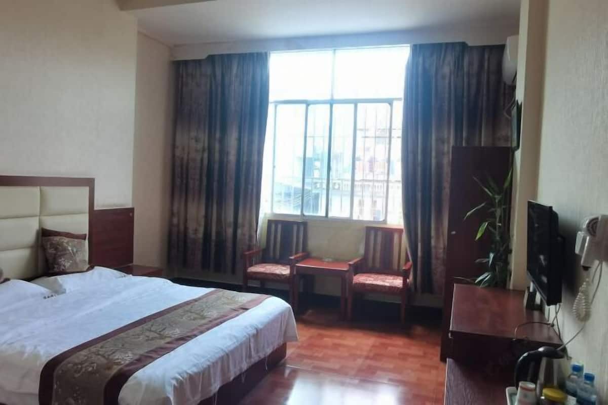 A hotel room with a large bed, a small desk, two chairs, a television, and a window with curtains.