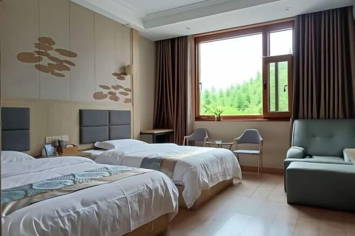 A hotel room with two beds, a desk, a chair, and a view of greenery through the window.