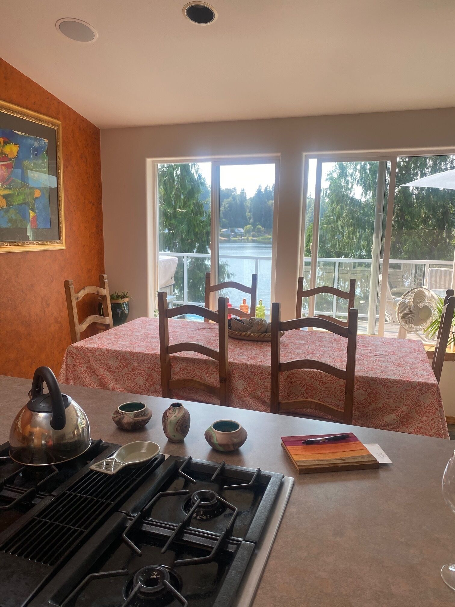 Une cuisine équipée d’une cuisinière à gaz, d’une bouilloire et d’un service à thé, une table à manger avec des chaises, et une grande fenêtre donnant sur un lac.