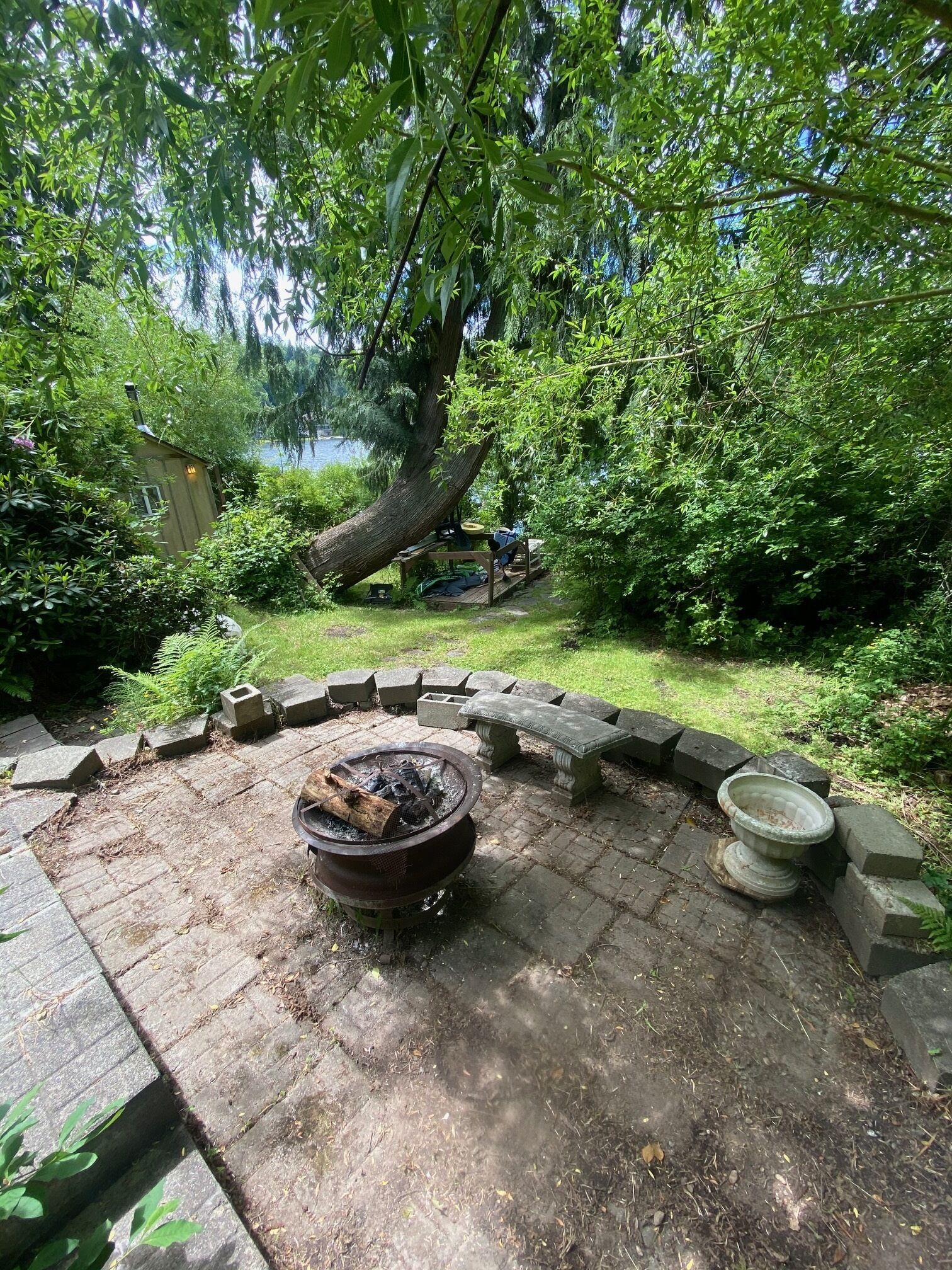 Un brasero rodeado de asientos de piedra, con un jardín exuberante y un árbol grande al fondo.