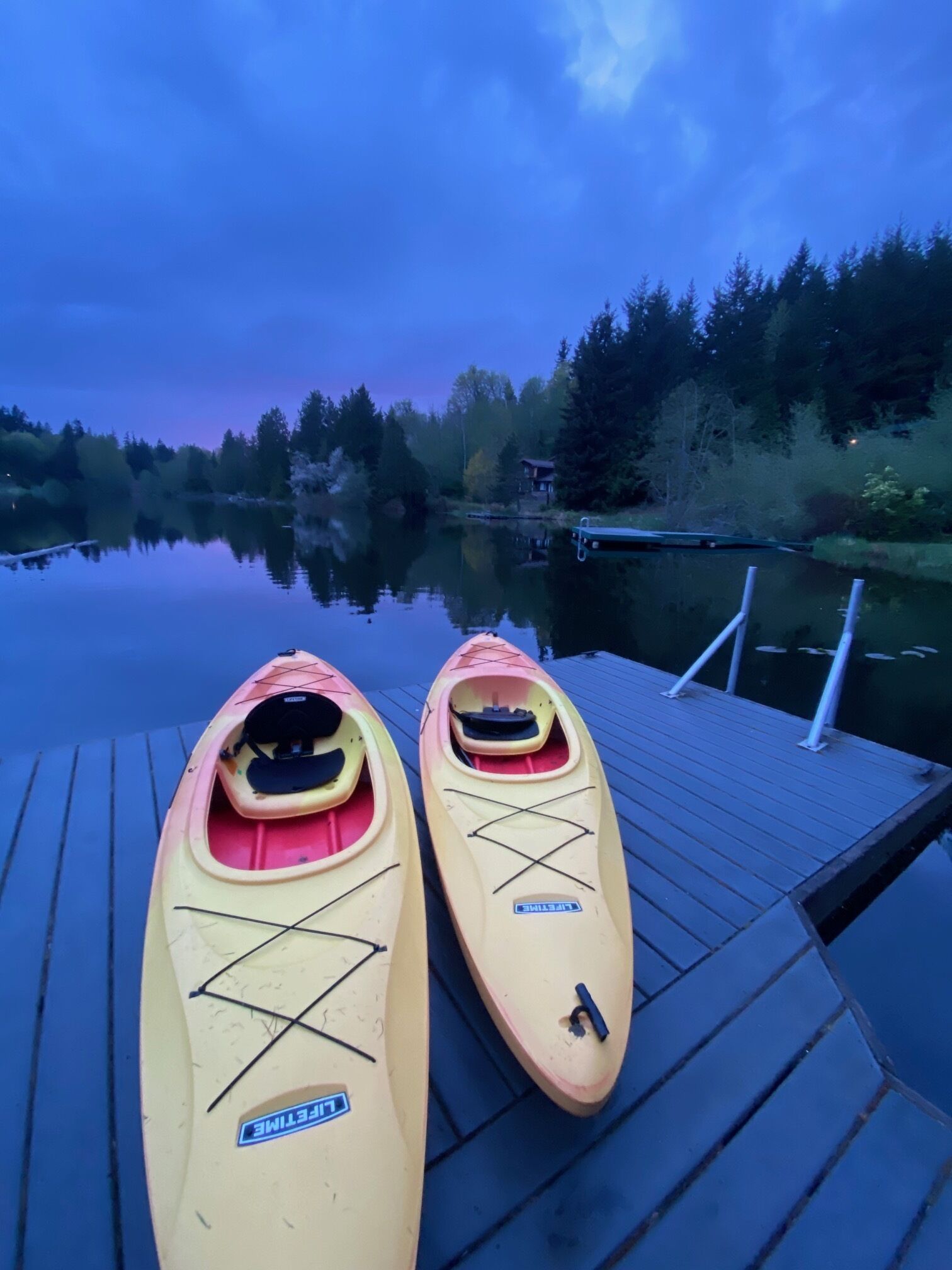 Two kayaks on a wooden dock with a calm lake and forest in the background.