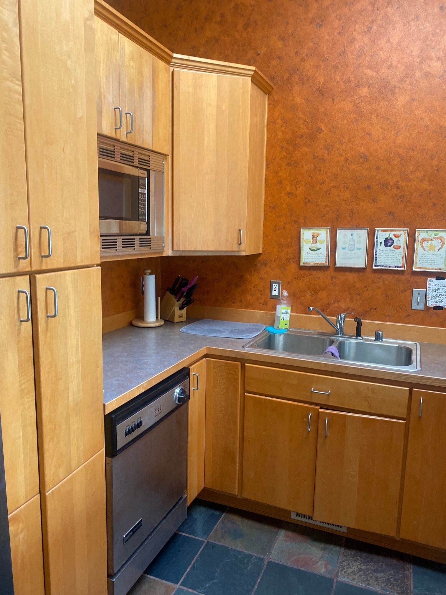 Una cocina compacta con armarios de madera, un microondas integrado, un lavavajillas y un fregadero. Hay cuatro diplomas enmarcados en la pared.