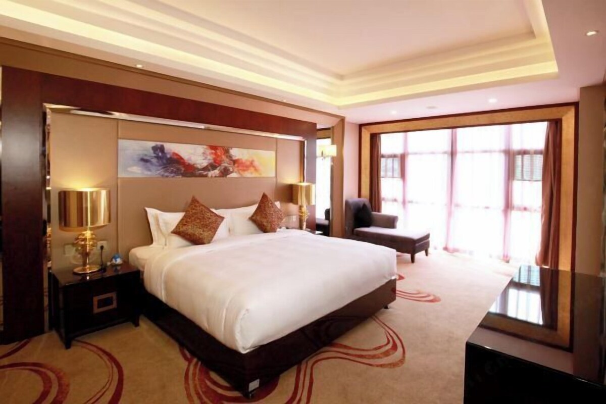 A large bed with white linens and multiple pillows, a bedside table with a lamp, a large window with curtains, and a modern ceiling design.
